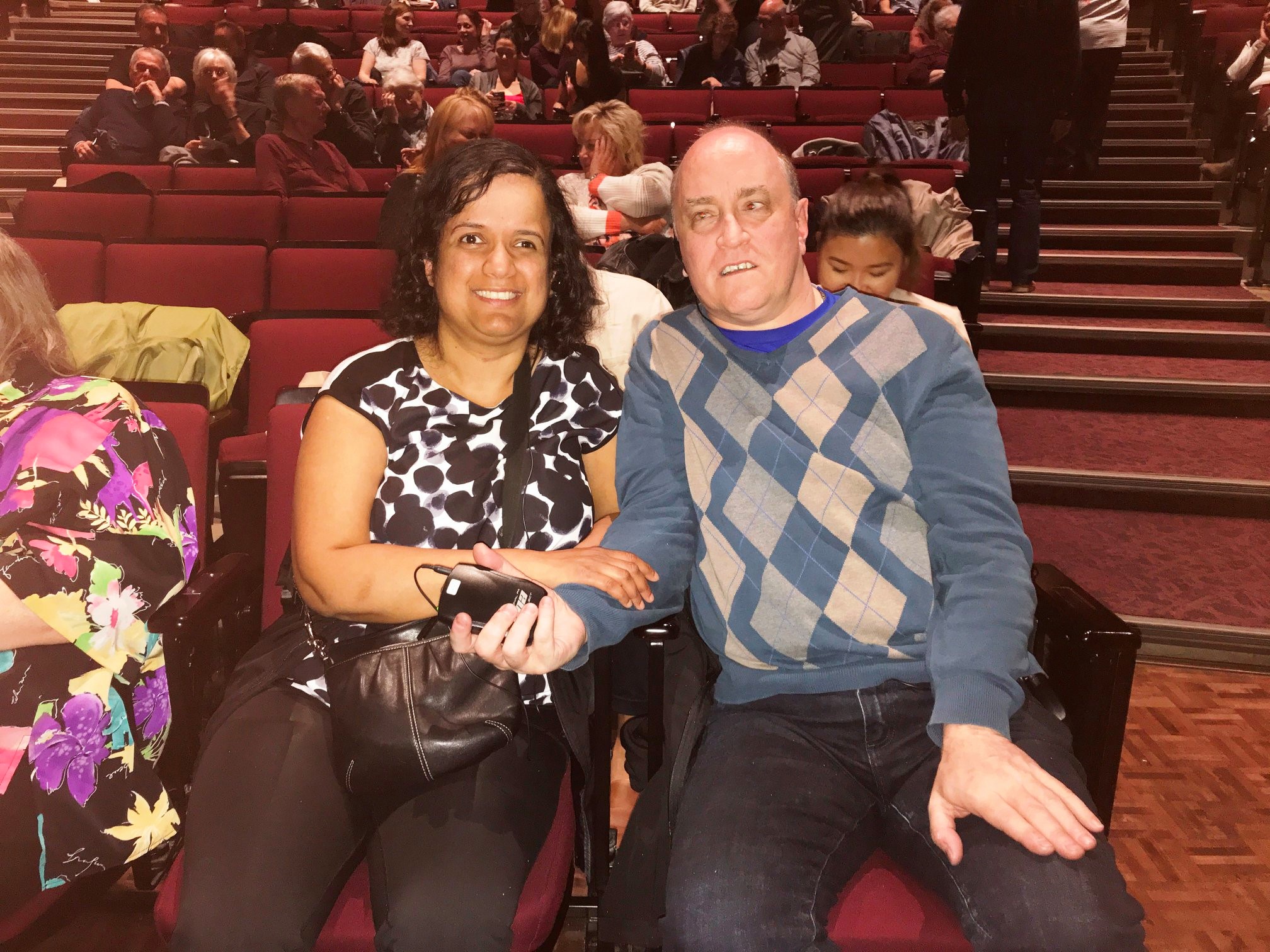 Kristy and Shawn sitting in the theatre, front row, with earpieces and transmitters for live audio description