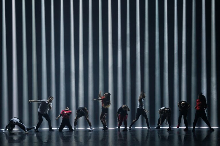 A black stage with a black background and 23 beams of light going straight down, making the back graphic look like jail bars or a bar code. They are at least 25 feet high based on the 10 dancers in front of them. Dancers wear red, black or white and appear to be battling something - some punch, some kneel.