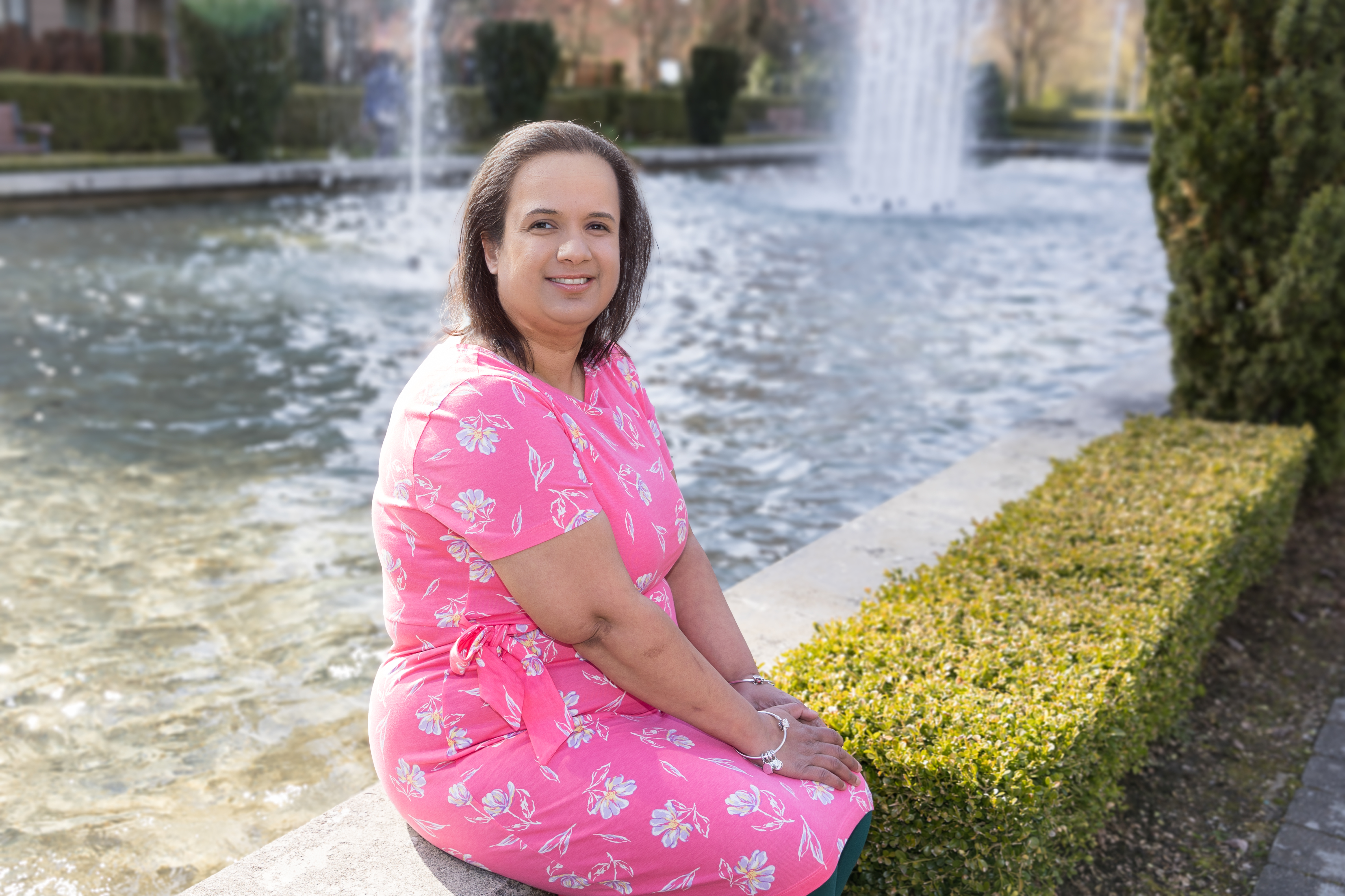 Kristy, in a red blouse over a black and white top, curly black hair framing her medium-brown skinned face, sitting on her patio with pink and blue hearts painted on the glass door behind her.