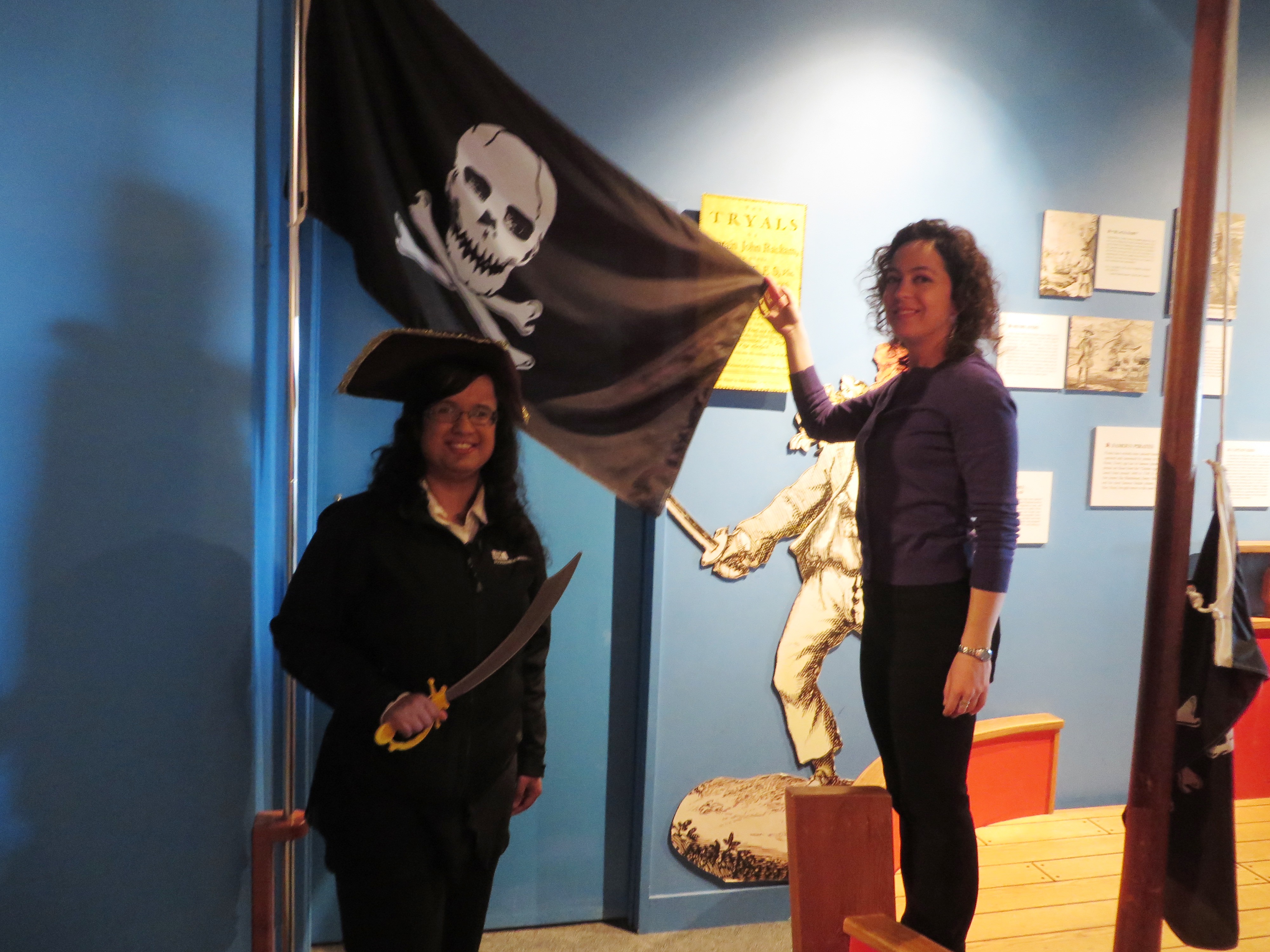 Kristy dressed as a pirate, standing in front of a jolly roger pirate flag
